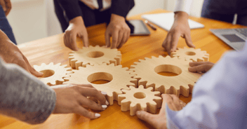 a closeup of a group of people's hands working to put a puzzle together. the puzzle is comprised of a set of large wooden gears. 